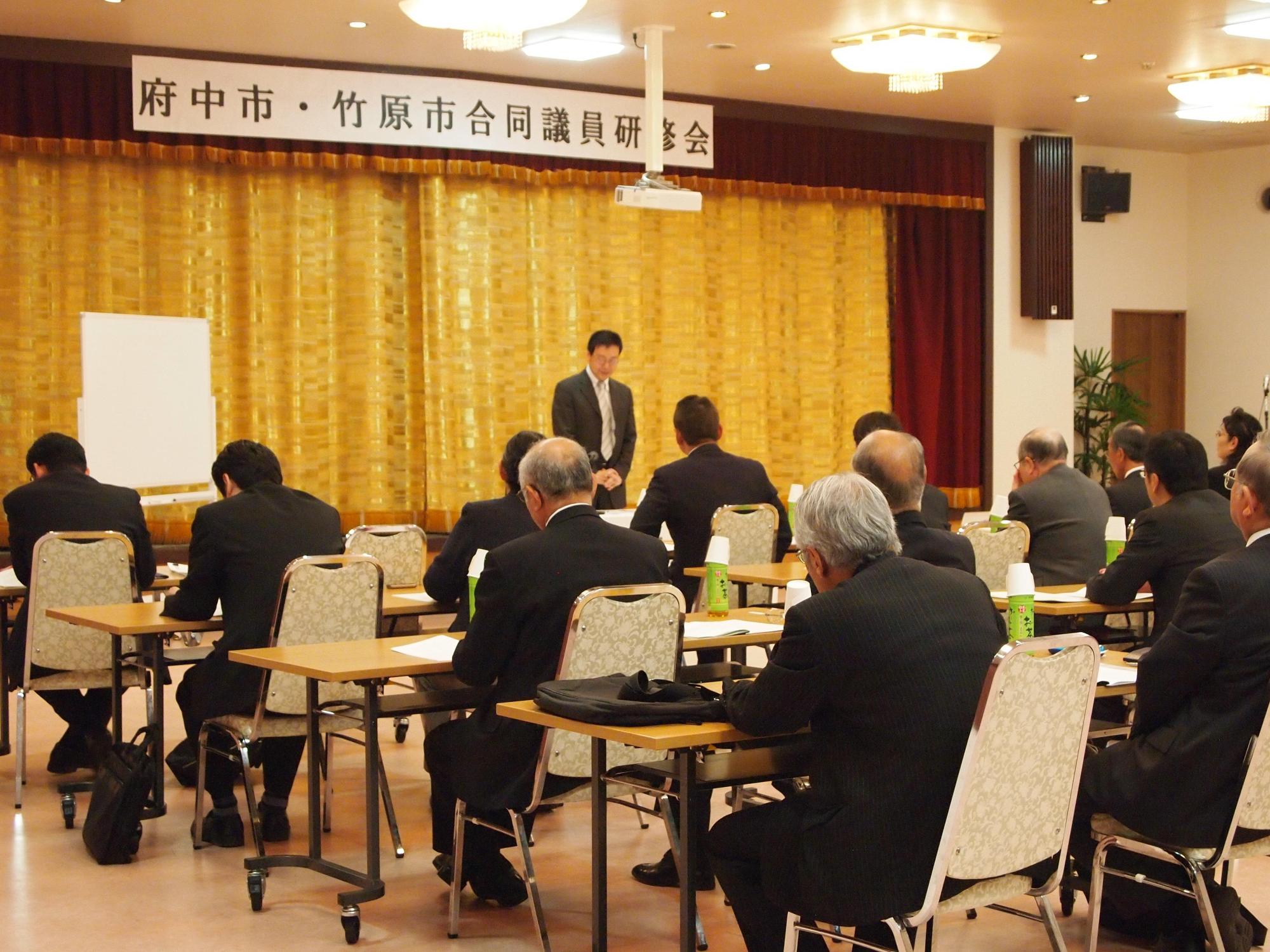 垂れ幕前で演説するスーツ姿の男性の前で、それぞれ設けられたテーブルに座り資料や話を聞く議員研修会の写真