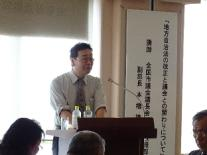 日が当たる背景で男性が講演をしている姿を撮影した写真