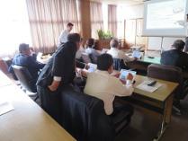 日光が差す室内でベージュ姿の男性がスーツを着た講師にタブレット操作を教わる様子を後ろから撮影した写真