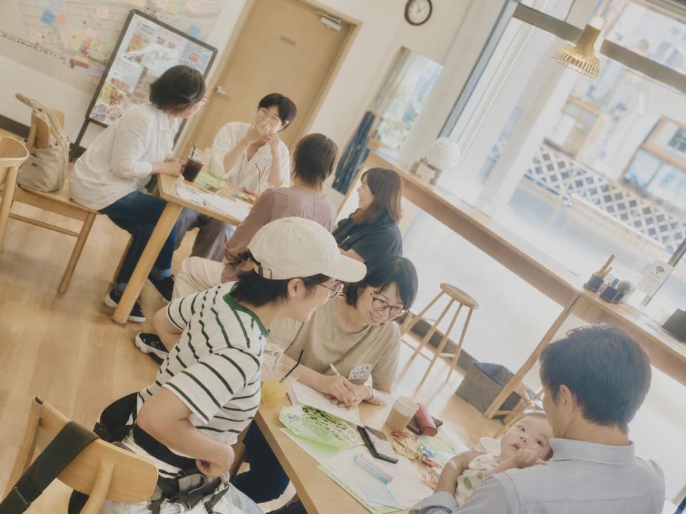 明るく木の温かみのあるコミュニティスペースで、赤ちゃんを抱く男性と女性たちが楽しそうに交流する様子の写真