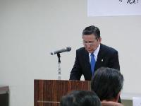 黒のスーツに青いネクタイをつけている市長が壇上でマイクに向かって発言している様子の写真