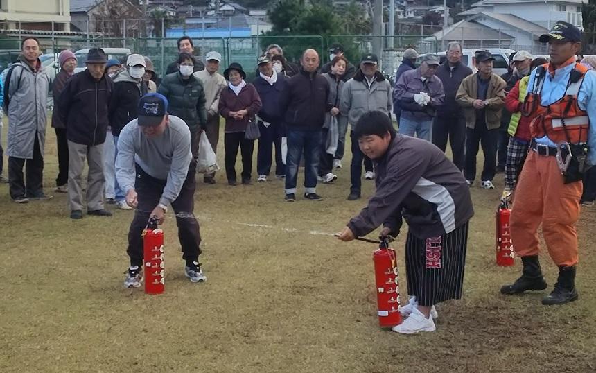 消火器のホースを手にしている2人の男性とそれを見ている防災訓練の参加者の写真