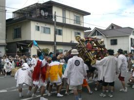 白い上着を羽織った人々が、道路の上で、神輿のようなものを持ち上げている写真