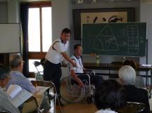 車椅子に乗っている人へのアプローチのしかたを説明している先生とその話を聞いている参加者の写真
