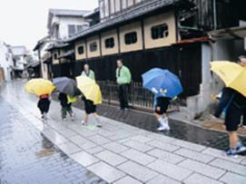 雨の中あいさつ運動をしている緑のジャンバー姿の大人たちと傘をさしている小学生たちの写真