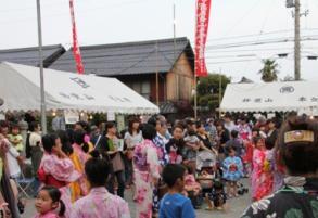 赤いのぼりが二つあり、白い屋根が二つあり、その周辺に集まる浴衣姿の人々の写真