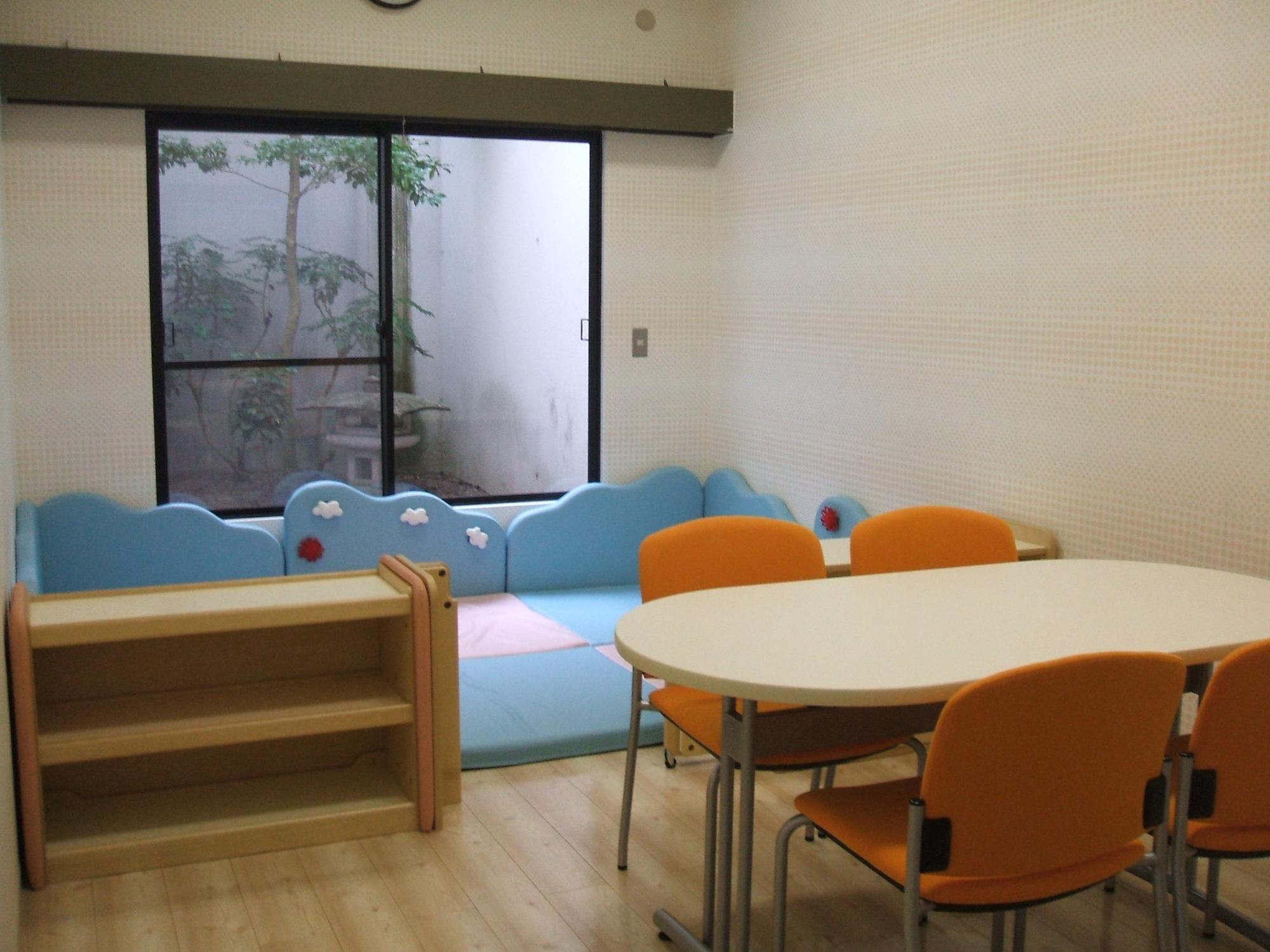 手前にオレンジ色の椅子と木製のテーブルが設置されており、奥には水色のマットのキッズスペースと靴箱が設置されているネウボラ相談室の写真