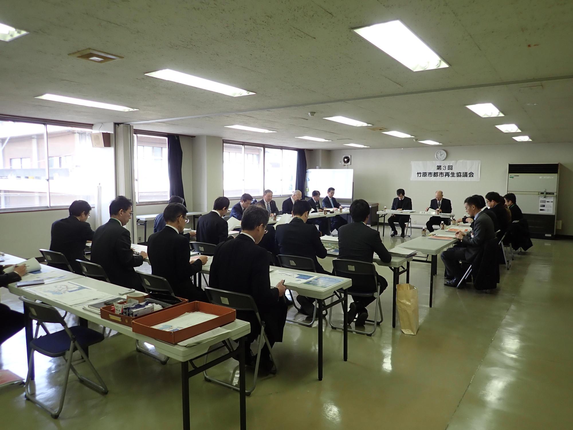 窓から日差しが差し込んでいる部屋で大勢が集まって第3回都市再生協議会を開催しているところを斜めから撮影した写真