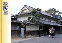 松阪邸 松が垣間見える瓦屋根の建物の傍を二人の人が歩いている写真