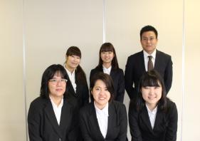 白い壁を背景に集まるスーツを着た男女6人の新入職員の写真