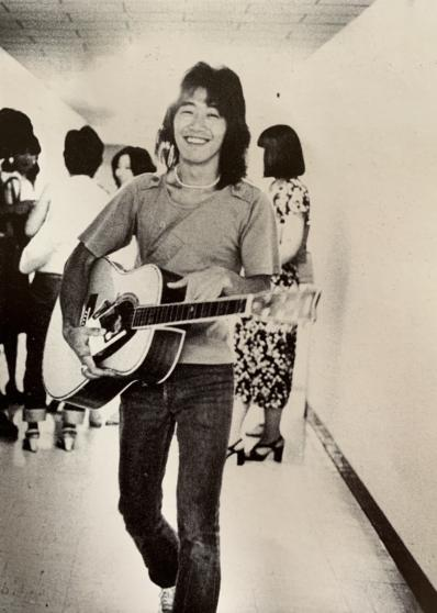 長髪でギターを抱え笑顔を見せている男性の白黒写真