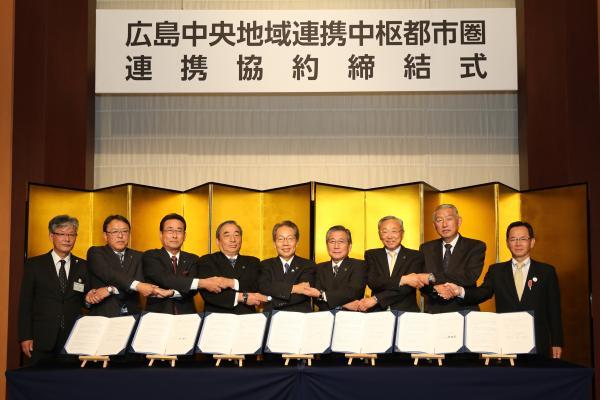 広島中央地域連携中枢都市圏連携協約締結式において9人の男性が7枚の連携協約書の後ろで横一列に並び手をつないでいる様子を撮影した写真